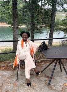 A woman sitting at a table near a lake, wearing a stylish cream outfit with an orange scarf, surrounded by trees.