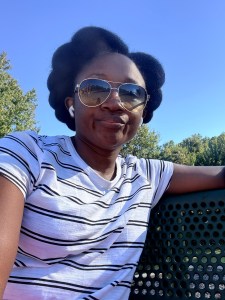 A young woman wearing sunglasses and a striped t-shirt, sitting on a bench outdoors with greenery in the background.